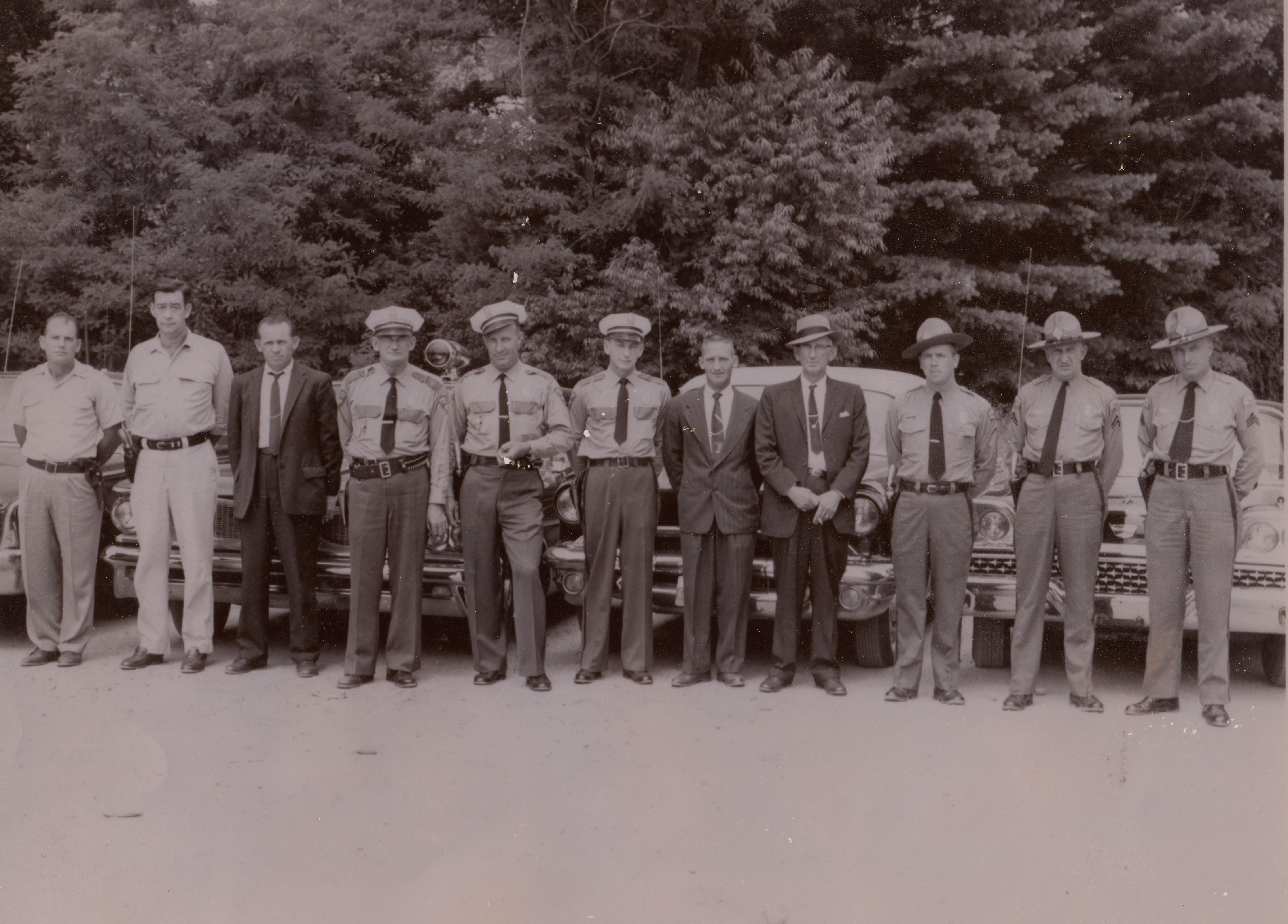 This photograph shows the Mitchell County Law Enforcement team about 1960. Pictured, left to right, are James Edwards and Joe Stewart, Mitchell County deputies; Sam Gouge, Mitchell County Sheriff; Jim Burleson, Spruce Pine Chief of Police; Ray Gunter and Landon “Cotton” Burleson, Spruce Pine Police Department officers; Herb Teague, Spruce Pine Police Department detective; Zeke Howell, Mitchell County Jailer; and North Carolina Highway Patrolmen Charles Radcliffe, R.E. Pipes, and Lee Lance. The automobiles, left to right, have been identified as a 1960 Ford, a 1960 Pontiac, a 1959 Chevrolet, and a 1959 Ford.